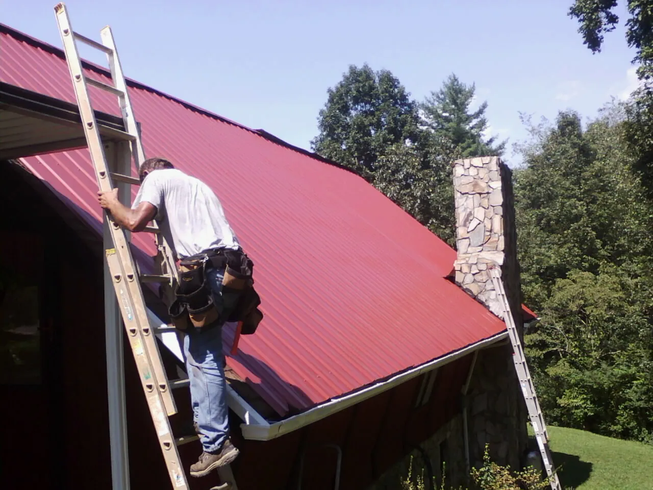 Roofer on a ladder beside a red metal roof and stone chimney — Soffit & Fascia Repair by Roofing Brothers in San Mateo