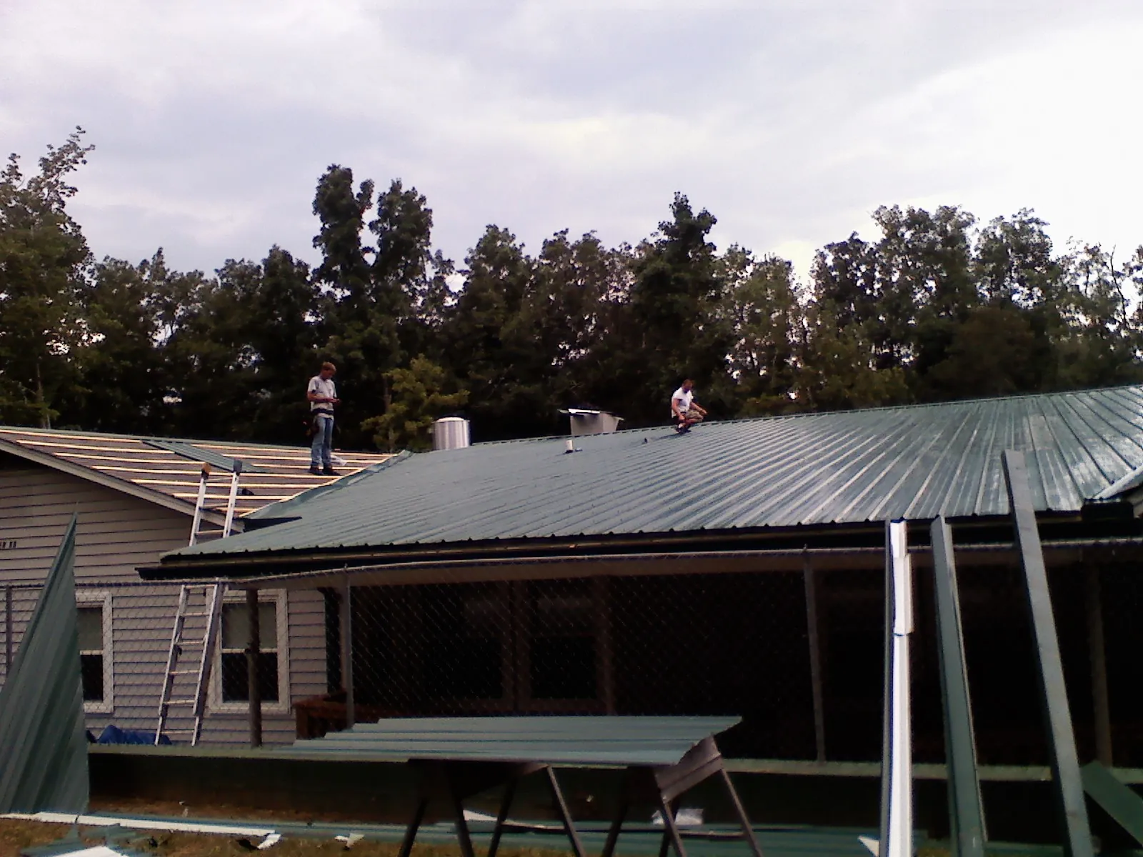 Two roofers installing green metal panels above a covered porch in San Mateo